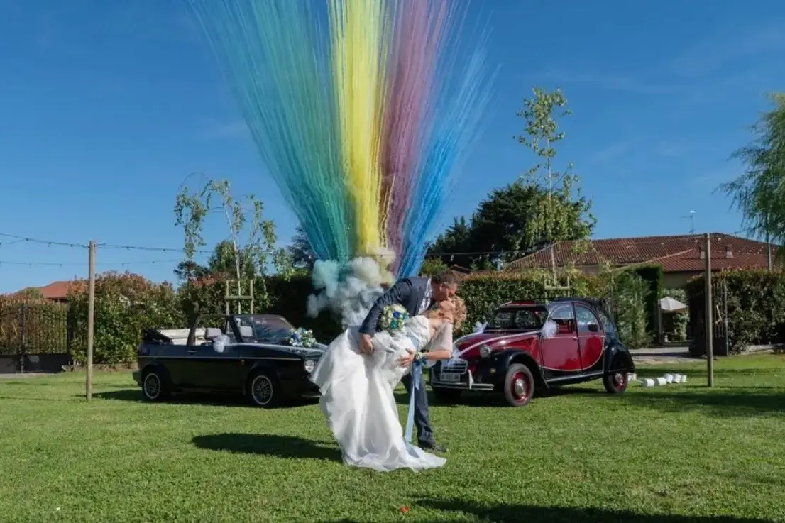 Sposi si baciano davanti a fumogeni colorati e auto d’epoca nel giardino