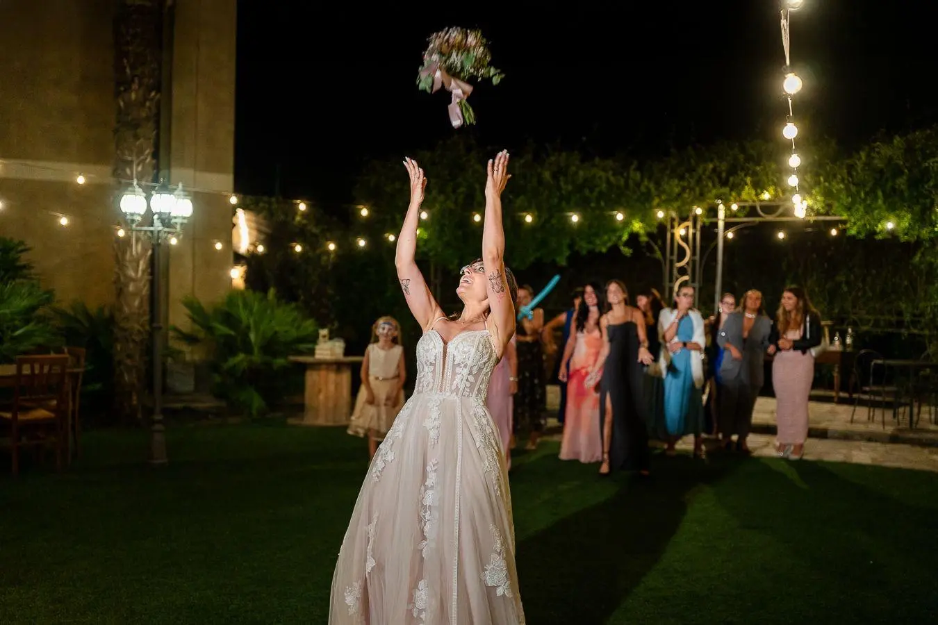 Sposa lancia il bouquet durante la festa serale nel giardino illuminato
