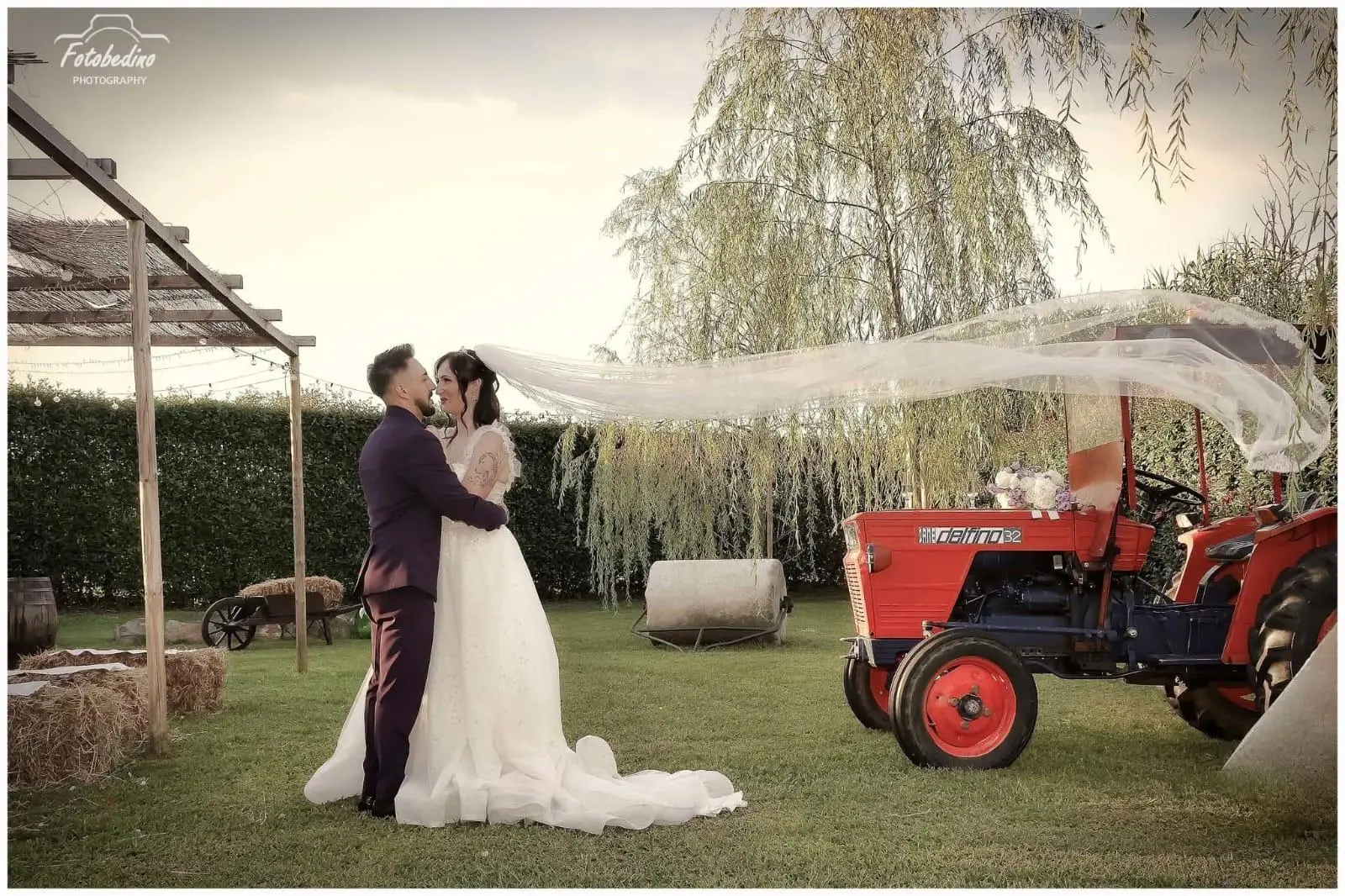 Sposi nel giardino accanto a un trattore rosso con il velo della sposa al vento